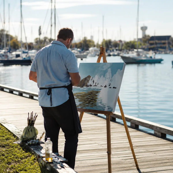 Peintre utilisant un chevalet en bois en plein air au bord d’un port, idéal pour la peinture extérieure.